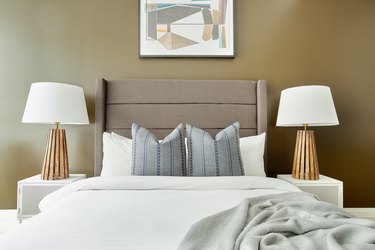 Modern bedroom with beige upholstered headboard, wood lamps, white nightstands, pillows, blanket, gold wall.